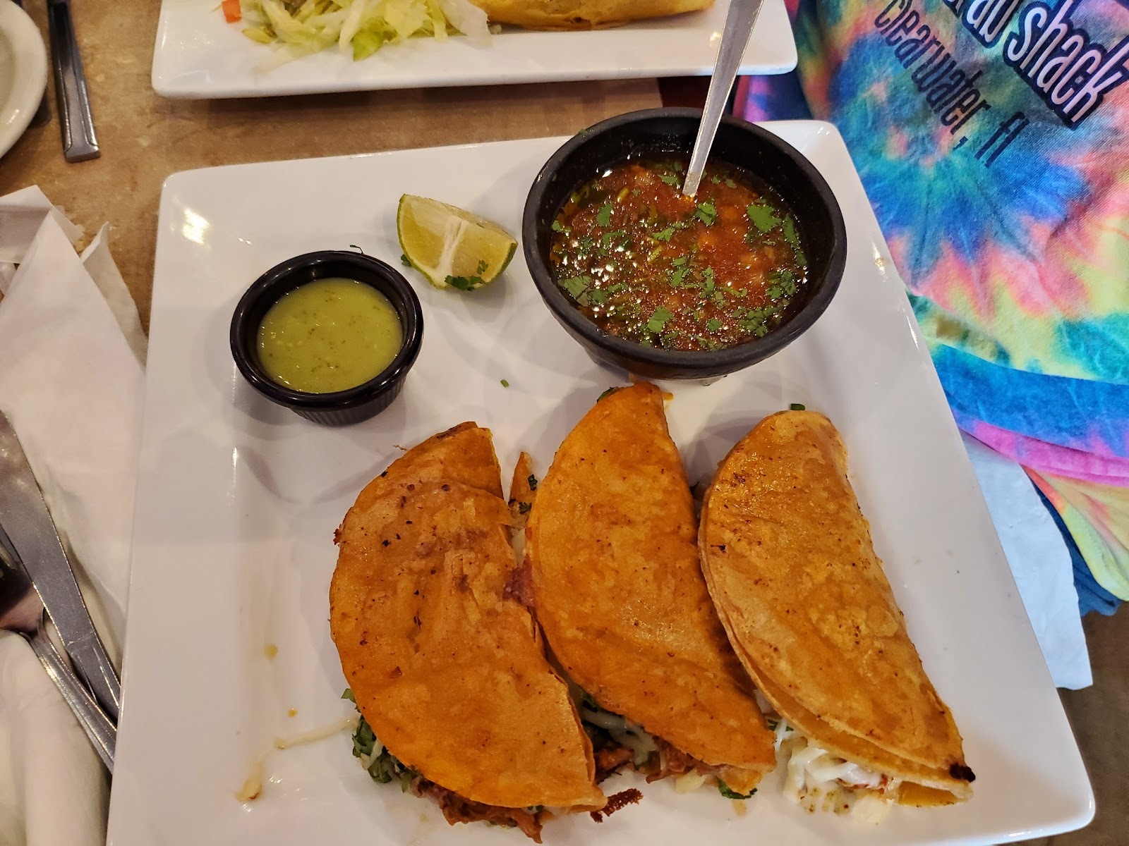 Three griddled quesabirria tacos on a white plate with a bowl of consomé, salsa verde and a lime wedge.